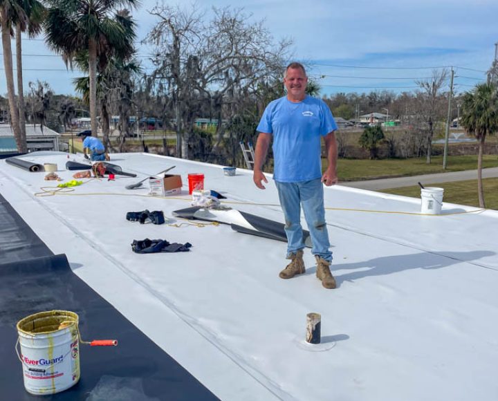 Roofer standing on a newly finished commercial flat roof, showcasing a completed roofing project with tools around, under a clear sky.